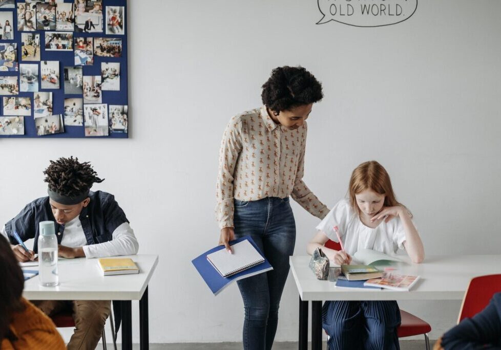 Adult standing beside a student seated at a desk in a classroom